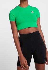 Vihreä lyhythihainen cropped t-paita, jossa on pieni logo; yhdistetty korkeavyötäröisiin mustiin pyöräilyshortseihin, sileä rakenne, tiukka muotoilu.