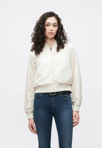 Bomber bunda krémové barvy se zipem vpředu, žebrovanými manžetami a pasem. Kombinováno s tmavě modrými úzkými džíny. Modelka má kudrnaté vlasy.