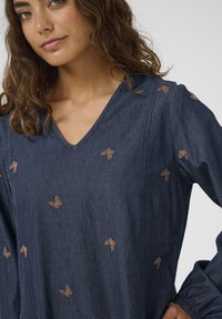 Denimbluse mit V-Ausschnitt, verziert mit kleinen bestickten braunen Schmetterlingen. Lange Ärmel mit elastischen Bündchen, Strukturstoff.