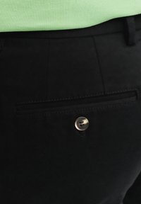 Czarne spodnie mają tylną kieszeń z zapięciem na guzik. Materiał jest gładki i nieco teksturowany, z widocznymi detalami szwów.