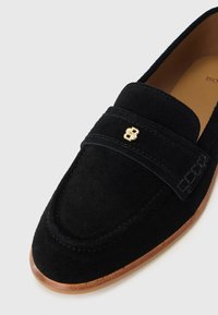 Mocasín de ante negro con punta redondeada, que presenta un emblema dorado en la correa y una suela de goma marrón claro. Parte superior texturizada con detalles de costura.