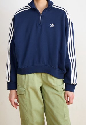 Osoba nosící námořnickou mikinu Adidas na čtvrtzip s bílými pruhy na rukávech a světle zelené kapsáčové kalhoty.