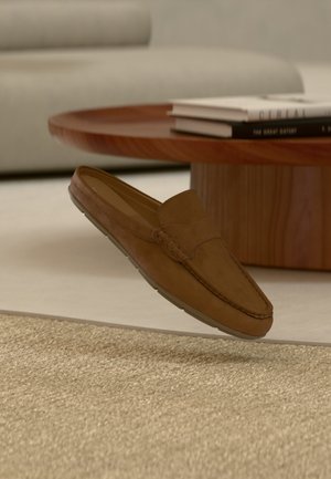 Rjavi usnjeni loafer lebdi nad bež tkanim preprogom, obkrožen z okroglo leseno mizo in zloženimi knjigami v mehko osvetljeni dnevni sobi.