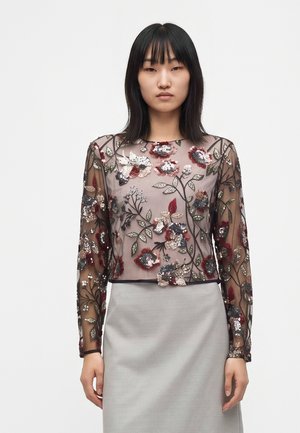 Femme portant un haut transparent à manches longues avec des sequins floraux rouges, noirs, argentés et verts, associé à une jupe gris clair sur fond blanc.