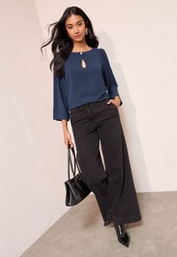 Marineblauwe blouse met een keyhole-detail, gecombineerd met hoge taille, wijd uitlopende zwarte jeans en zwarte puntige laarzen. Accessoires omvatten een zwarte handtas.