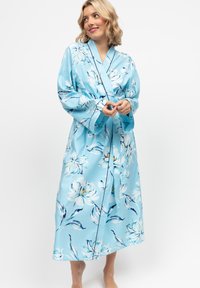 Donna che indossa una vestaglia di satin a fiori blu chiaro, in piedi a piedi nudi contro uno sfondo bianco semplice, guarda di lato sorridendo.