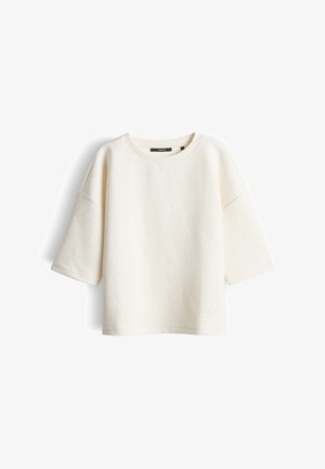 Crèmekleurig topje met korte mouwen, voorzien van een subtiel reliëf met bloemenpatroon en een ronde halslijn, weergegeven op een witte achtergrond.