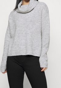 Světle šedý pletený svetr s vysokým límcem, uvolněným střihem a kleslým ramenem. Hladký žebrovaný vzor a lemování. Nosený přes černé kalhoty.