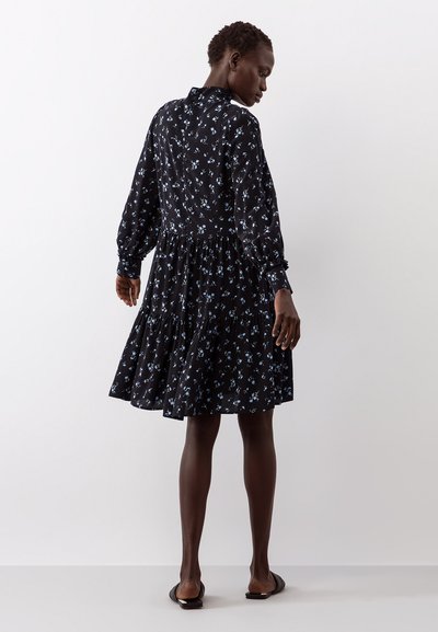 Vestido preto com padrão floral azul, mangas compridas bufantes, saia evasê e um design em camadas. Vista traseira, tecido suave, detalhe na gola.