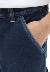 Jean en denim bleu foncé avec un design traditionnel à cinq poches, présentant des coutures visibles le long des coutures et une main dans la poche avant.