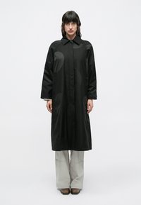 Femeie cu păr închis la culoare, purtând o haină lungă neagră cu modele circulare subtile, pantaloni gri deschis și pantofi maro, stând în fața unui fundal simplu.