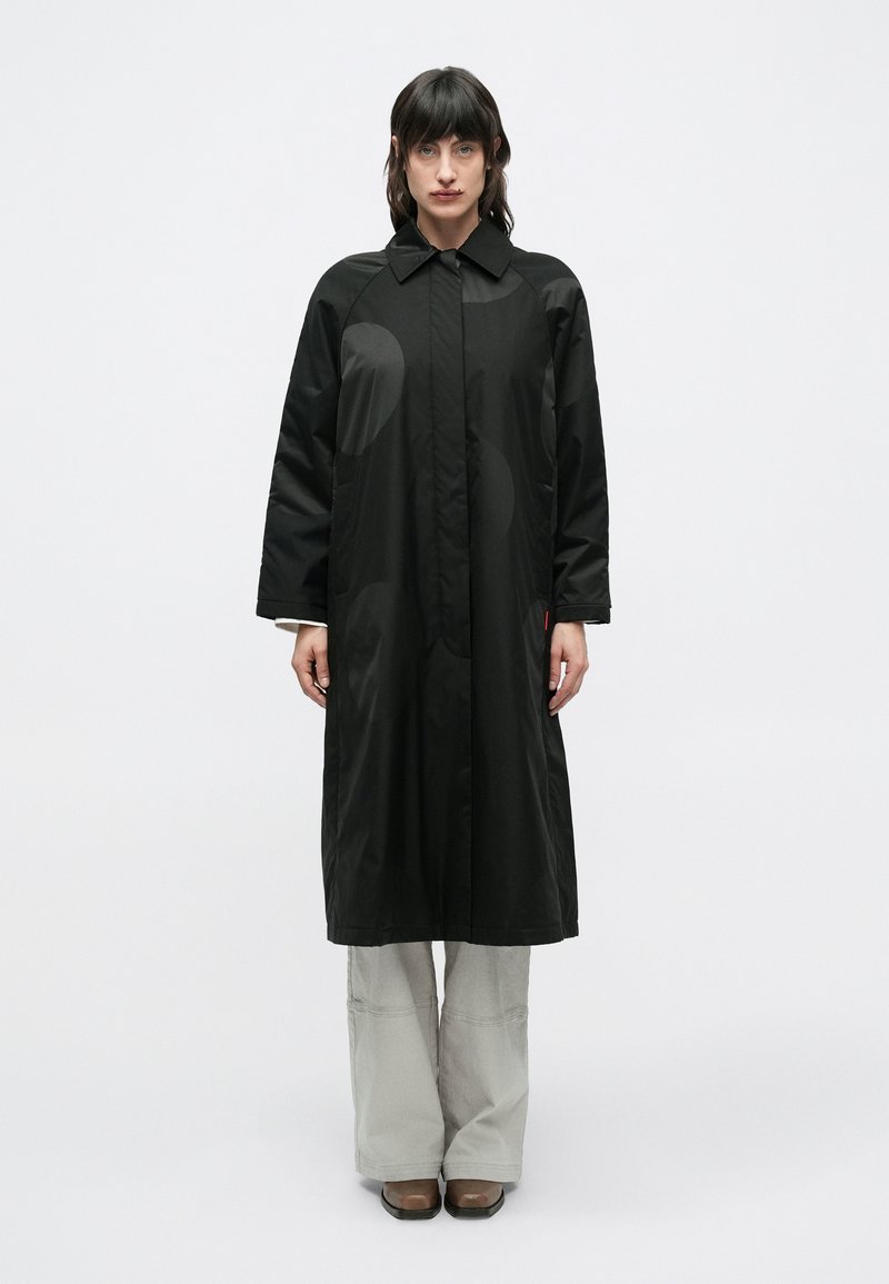 Femeie cu păr închis la culoare, purtând o haină lungă neagră cu modele circulare subtile, pantaloni gri deschis și pantofi maro, stând în fața unui fundal simplu.