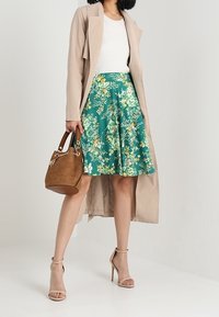 Una mujer se encuentra de pie con una falda verde floral y un abrigo beige, sosteniendo un bolso marrón, mostrando un atuendo elegante con tacones de cinta.