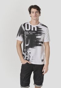 T-shirt à manches courtes avec un design abstrait en noir et blanc, présentant un texte audacieux et des motifs de coups de pinceau sur le devant, fabriqué en coton.