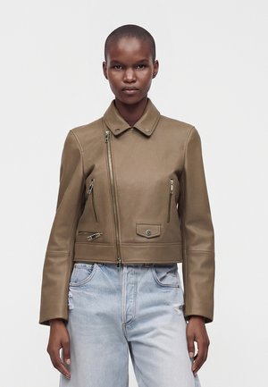 Vrouw met kort haar die een bruine leren jas met meerdere ritsen draagt en lichtblauwe, hoog getailleerde jeans met lichte slijtage.
