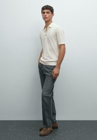 Ung man står klädd i en krämfärgad kortärmad polo, grå jeans och bruna mockaloafers mot en enkel ljusgrå vägg.