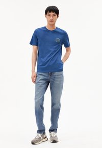 T-shirt di cotone blu con maniche corte, scollo rotondo e grafica di un sorriso giallo sul petto. Abbinata a jeans azzurri chiari e sneakers.