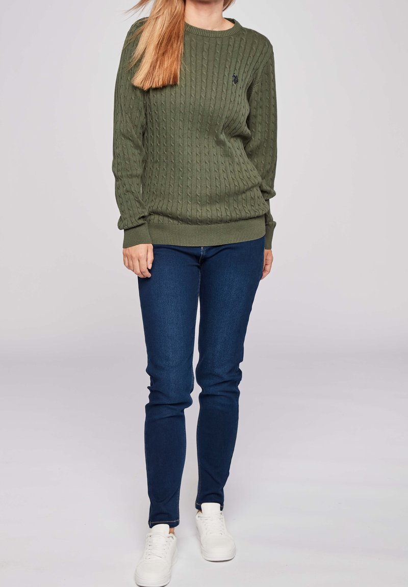 Femme portant un pull torsadé vert olive, un jean bleu foncé et des baskets blanches, debout devant un fond clair uni.