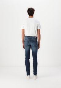 Jeans de denim ajustados en lavado azul oscuro, con bolsillos traseros y una cinturilla con logo. Combinados con una camiseta blanca sencilla y zapatos blancos.