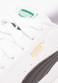 Baskets en cuir blanc avec des accents noirs, arborant un logo doré "PUMA BASKET" et une languette texturée avec une étiquette verte.