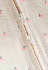 Lähikuva beigevärisestä, poimutetusta kankaasta, jossa on vaaleanpunaisia kirsikkakuoseja ja keskellä osittain vetoketjulla avattu valkoinen kaksinkertainen vetoketju.