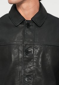 Jaqueta de couro preta com fecho frontal de botões, grande gola e superfície texturizada. Apresenta quatro botões pretos e detalhes em costura contrastante.