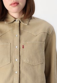 Bež kravljasti jeans s pointed ovratnikom, dvema sprednjima žepekoma in gumbi na pritisak. Ima rdečo oznako logotipa na levem žepu.