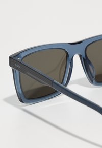 Óculos de sol azuis com uma armação de plástico brilhante, lentes escuras e hastes pretas texturizadas com um logótipo subtil de lado.