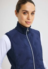 Marinblå pufferväst med hög krage, med dragkedjeslutning och diskret broderi. Slät textur med en figurnära design, lager-på-lager över en vit topp.