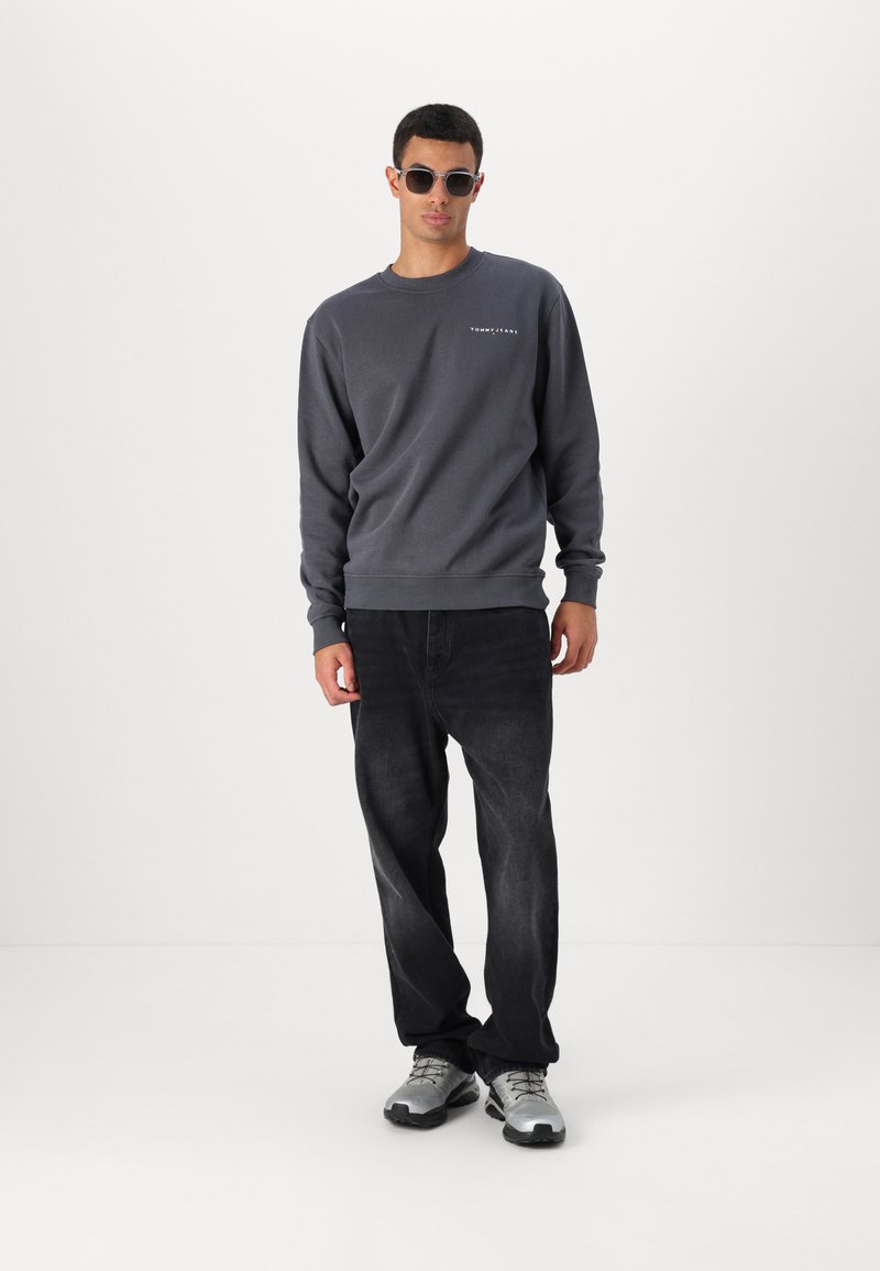 Felpa grigia con scollo rotondo e logo, abbinata a jeans in denim neri e scarpe da ginnastica grigie. Il modello indossa occhiali da sole.