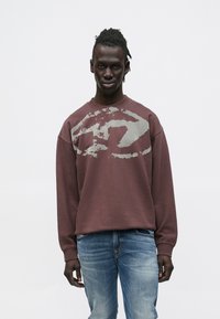 Moški temne polti z dreadlocki, oblečen v bordo rjavo jopico z veliko sivo abstraktno grafiko in izpranimi modrimi kavbojkami, stoji na belem ozadju.