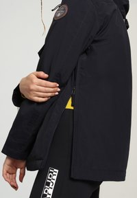 Giacca da esterno blu navy con una patch sulla manica, zip laterali e una vestibilità comoda, abbinata a un top giallo vivo sotto.