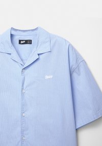 Camicia a maniche corte azzurro chiaro con bottoni, caratterizzata da sottili righe verticali e un piccolo logo bianco nell'area della tasca sul lato sinistro del petto. Design con collo piatto.