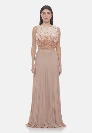 Frau trägt ein ärmelloses, zweifarbiges beiges Kleid mit einem strukturierten floralen Oberteil und langem plissiertem Rock, steht vor einem schlichten Hintergrund.