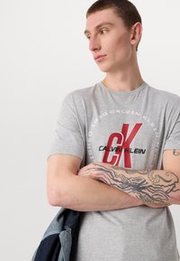 Gray cotton t-shirt featuring a circular graphic with "Calvin Klein" and "Established 1958" text; bold red "CK" logo at center.
