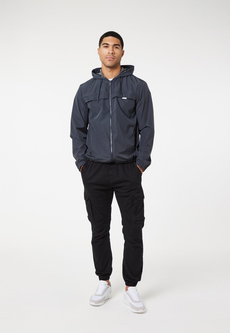 Marineblauwe windbreaker met een ritssluiting aan de voorkant en een capuchon, gecombineerd met zwarte cargo-broeken met meerdere zakken en witte sneakers.