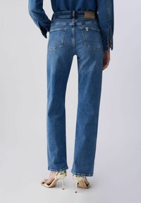 Naine, kes kannab sirge säärega siniseid teksapükse ja kuldseid kontsakingi, nähtud tagantpoolt tavalise tausta ees.