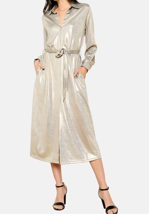 Žena má na sobě zlaté lesklé košilové šaty s dlouhým rukávem a páskem, s kapsami, doplněné černými sandály na vysokém podpatku s páskem kolem kotníku.