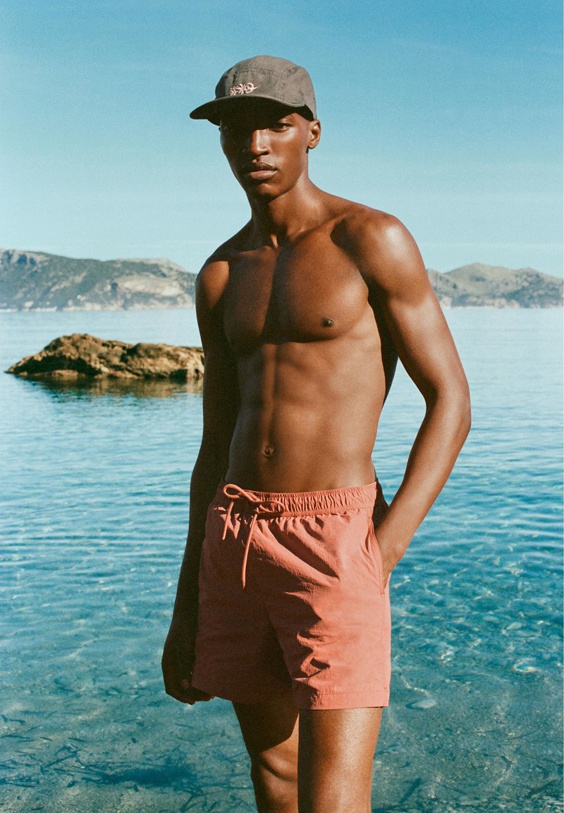 Homme torse nu portant une casquette et un short de bain corail, debout dans une eau claire peu profonde avec des îles rocheuses en arrière-plan.
