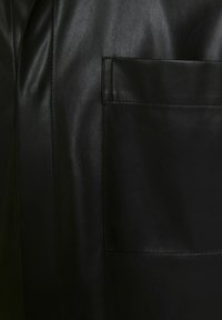 Svart skinnjacka med en slät yta, en rektangulär ficka fram och synlig söm längs sömmarna.