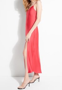 Rode satijnen maxi-jurk met een V-hals, dunne bandjes en een zijsplit. Gemaakt in combinatie met doorzichtige haksandalen. Glad textuur en aansluitend ontwerp.