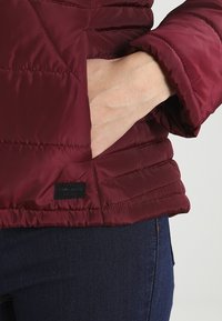 Bourgognefarvet dynejakke med tekstureret finish, der har en sidelomme og et sort mærke med "Vero Moda" ved kanten.