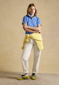 Žena stojaca so založenými rukami, oblečená v modrom polo tričku, bielych nohaviciach, žltom svetri previazanom okolo pása a žltých teniskách s textúrovanými ponožkami.