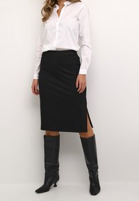 Schwarzer Bleistiftrock mit seitlichem Schlitz, kombiniert mit einem weißen Hemd mit Knöpfen. Modell trägt kniehohe schwarze Lederstiefel.