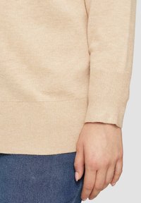 Beige gestrickter Pullover mit gerippten Bündchen und Saum, mit einer weichen Textur. Teilweise sichtbare Hand, die auf einer dunkelblauen Jeans ruht.