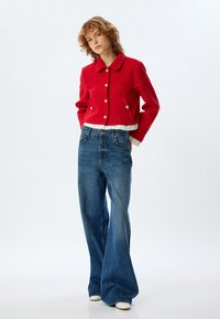 Rote, verkürzte Jacke mit weißem Innenfutter und goldenen Knöpfen, kombiniert mit hochgeschnittenen blauen Jeans im Bootcut-Stil und weißen Schuhen.