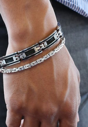 Twee armbanden om een pols: één zwart en zilver, met in elkaar grijpende schakels, en een zilveren gevlochten ontwerp. Gladde en gestructureerde oppervlakken.
