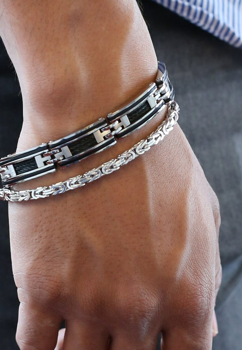 Twee armbanden om een pols: één zwart en zilver, met in elkaar grijpende schakels, en een zilveren gevlochten ontwerp. Gladde en gestructureerde oppervlakken.