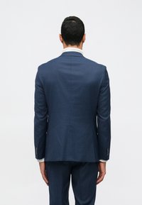 Ναυτικό μπλε κοστούμι με κομψή εφαρμογή, που διαθέτει απαλή υφή και διακριτό καρό σχέδιο. Μοναδική αεροξέστης στο πίσω μέρος, με λευκό πουκάμισο από κάτω.