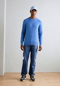 Hellblauer Strickpullover, blaue Jeans und schwarze Sneakers mit weißen und grauen Akzenten. Die Figur trägt eine hellblaue Kappe. Schlichter Hintergrund.